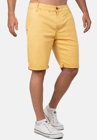 Geel katoenen shorts met opgerolde zoom, voorzien van rode accenten en zijzakken. Combinatie met witte sneakers en zichtbare enkel.