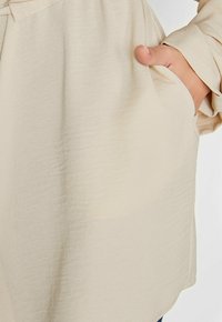 Beige Bluse mit strukturiertem Finish, entspannter Passform und Seitentaschen. Die Ärmel sind an den Handgelenken leicht gerafft.