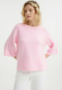 Maglione rosa, morbido, con scollatura a girocollo, maniche raglan corte e un fit rilassato. Abbinato a pantaloni bianchi aderenti.