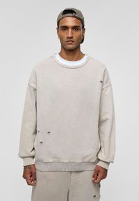 Lichtbeige sweatshirt met een ontspannen pasvorm, zichtbare versleten details, rib gebreide manchetten en een ronde hals, gedragen over een witte t-shirt.
