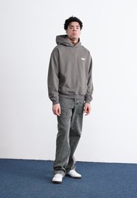 Grå hættetrøje med en frontlomme og logo, parret med slidte grå jeans og hvide sneakers. Ensfarvet baggrund og blåt tæppe.