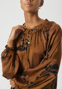 Brun blommig blus med en samlad halsringning, långa ärmar och knappdetaljer i guld och svart. Tillverkad av mjukt, lätt tyg.