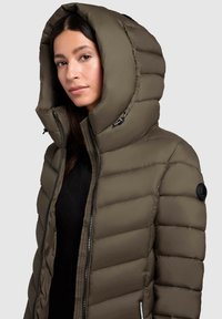 Olīvu zaļš pūkains mētelis ar augstu, polsterētu kapuci, ar rāvējslēdzēju un horizontālu pundurkociņu. Ietver apaļu logotipa uzlīmi uz piedurknes.