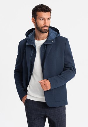 Veste bleu marine avec une capuche amovible, fermeture par boutons, deux poches et une texture lisse. Portée sur un pull blanc et un pantalon à carreaux.