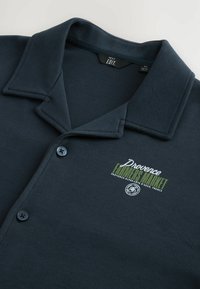 Marineblå polo skjorte fremstillet af blødt stof, med knaplukning, flad krave og broderet "Provence Farmers Market" logo.