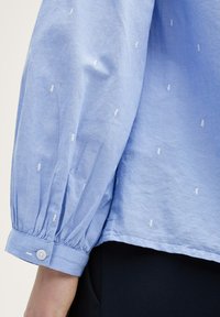 Blouse bleu clair avec des manches longues et bouffantes. Présente de petits détails brodés blancs, des poignets froncés et une fermeture à boutons. Tissu en coton.