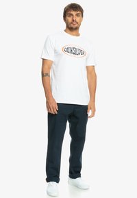 T-shirt en coton blanc avec un logo Quiksilver noir et orange. Associée à un pantalon bleu marine et des baskets blanches. Design simple et décontracté.
