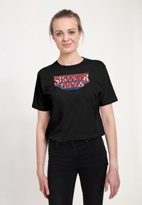 Camiseta negra de algodón con un colorido logo de "Stranger Things" en rojo y azul, destacado en la parte frontal. Mangas cortas, cuello redondo.