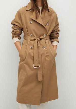 Femme portant un trench-coat croisé couleur beige avec ceinture à la taille et manches retroussées, par-dessus un pull crème et un pantalon blanc.