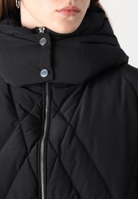 Crna podstavljena jakna s visokim ovratnikom i velikim kapuljačom, kviltani uzorak, srebrni patentni zatvarač i detalji s dugmetima.