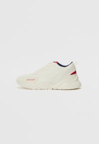 Sneaker in cream with textured leather and mesh, featuring red and navy accents. Rounded toe, lace-up design, and a thick rubber sole.