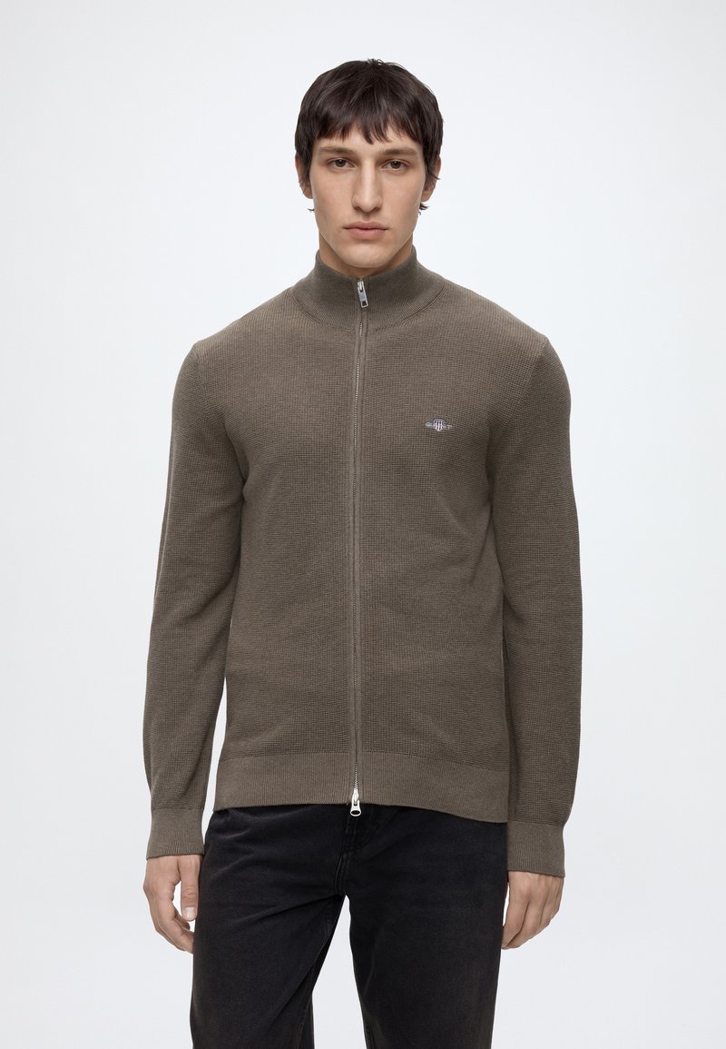 Hombre que lleva un jersey de punto marrón con cremallera y un pequeño logo en el pecho y pantalones negros, de pie frente a un fondo blanco liso.
