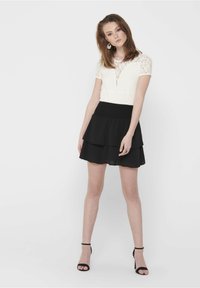 Weißes Spitzentop mit tiefem Ausschnitt, kombiniert mit einem schwarzen, geschichteten Chiffonrock. Das Outfit verfügt über einen figurbetonten Bund und schwarze Riemchen-High Heels.