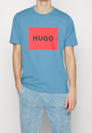 Homme portant un t-shirt bleu à manches courtes avec un carré rouge affichant le mot « HUGO » en lettres noires sur la poitrine.