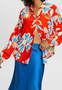 Chemise à boutons rouge à motifs floraux bleus et jaunes, manches longues, coupe décontractée, associée à une jupe en satin bleue.