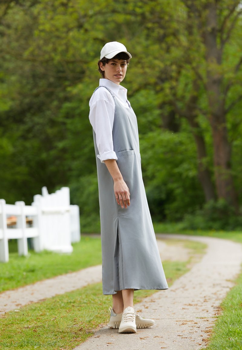 Abito midi grigio con spacchi laterali, indossato sopra una camicia bianca con bottoni. Completato con sneakers chiare e un cappellino bianco. Sfondo verde.