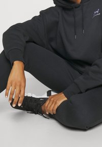 Felpa nera con cappuccio e lacci, indossata con leggings neri e scarpe da ginnastica nere. Presenta un logo sulla felpa. Tessuto dall'aspetto liscio.