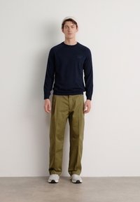 Mornarska majica z dolgimi rokavi in okroglim izrezom, v kombinaciji z olivno zelenimi hlačami. Model nosi sive športne copate. Preprost, funkcionalen dizajn na nevtralni podlagi.