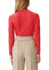 Vrouw in een rode lange mouwen coltrui, gestoken in hoog aangesloten beige broek met een riem, gezien van achteren.