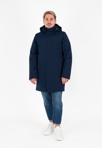 Parka impermeabile blu navale con colletto alto, zip frontale e cappuccio regolabile. Abbinato a jeans blu casual e sneakers bianche.