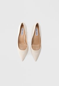 Μπεζ γοβάκια μυτερής μύτης με λείο συνθετικό επάνω μέρος, γυμνό πάτο και διακριτικό λογότυπο στο τόξο. Μινιμαλιστικός σχεδιασμός, χωρίς εμφανή μεταλλικά στοιχεία.