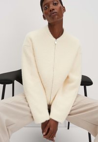 Gestructureerde crèmekleurige cardigan met een hoge kraag en een ritssluiting aan de voorkant, met brede mouwen en een zachte, donzige stof. Model zit en draagt beige broeken.