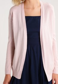 Ljusrosa stickad cardigan med långa ärmar, öppen front och ribbade muddar, bärs över en mörkblå klänning. Mjuk textur, klassisk design.