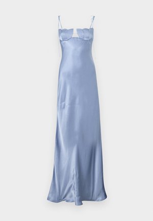 Femme portant une longue robe en satin bleu clair avec fines bretelles et un bustier festonné, debout les mains dans le dos sur un fond uni.
