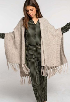 Pañuelo de lana beige claro con bordes con flecos, usado sobre una camisa a cuadros verde oscura y pantalones a juego. Textura suave, diseño holgado.