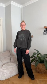 Tumehall pusa graafilise mustriga, oversize lõikega, kombineeritud lahtiste mustade pükste ja mustade jalanõudega. Ruumi iseloomustavad pehmed beežid mööbliesemed ja taimed.