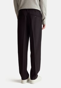 Černé široké kalhoty s hladkou texturou, se zadními kapsami na knoflíky, v kombinaci se šedým dlouhým rukávem a bílými teniskami.