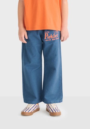 Child wearing blue wide-leg pants with orange text "Pickles" on left thigh, orange shirt, and striped sneakers with gum soles.