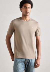 Νέος άνδρας με σγουρά μαλλιά, φορώντας μπεζ κοντομάνικο T-shirt με μικρό λευκό λογότυπο και ανοιχτόχρωμο μπλε τζιν, στέκεται μπροστά σε μονόχρωμο φόντο.