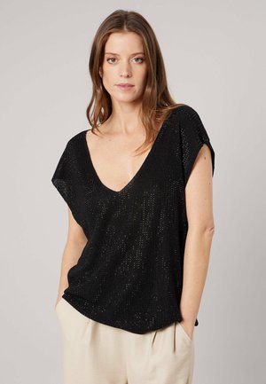 Femme aux cheveux châtain clair portant un haut noir en maille avec col en V et manches courtes, un pantalon beige, les mains dans les poches, sur un fond uni.