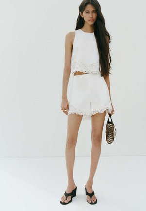 Femme aux longs cheveux foncés portant un haut blanc sans manches et un short avec des détails en dentelle, tenant un petit sac tissé noir et beige.