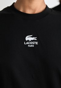 Schwarzes Rundhals-Shirt mit weißem Lacoste-Logo, das ein Krokodil und den Text "LACOSTE PARIS" auf der Brust zeigt.