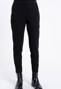 Czarne, dopasowane spodnie z detalami w postaci złotych zamków błyskawicznych w pasie, noszone z błyszczącymi czarnymi botkami, przedstawione od pasa do stóp na białym tle.