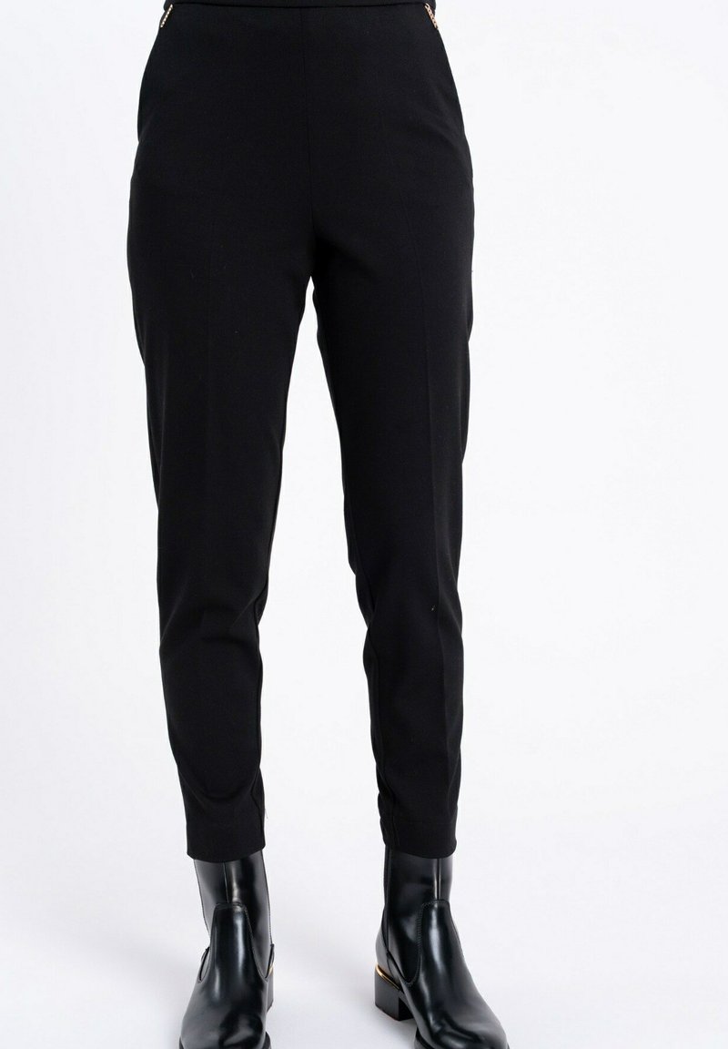 Czarne, dopasowane spodnie z detalami w postaci złotych zamków błyskawicznych w pasie, noszone z błyszczącymi czarnymi botkami, przedstawione od pasa do stóp na białym tle.