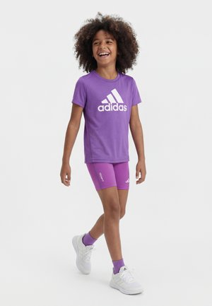 Enfant aux cheveux bouclés marchant, portant un t-shirt Adidas violet, un short Techfit assorti, des chaussettes violettes et des baskets blanches, souriant et regardant vers la droite.