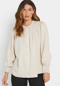 Blouse crème à manches longues avec un devant boutonné, des accents froncés et des poignets smockés. Fabriquée en tissu léger avec une texture douce.
