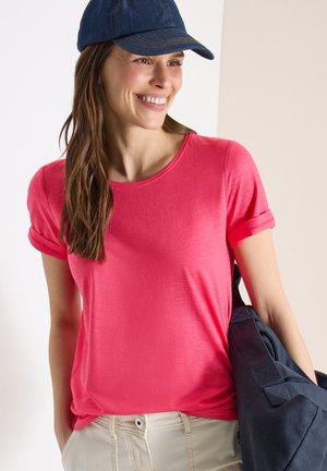 Femme souriante portant un t-shirt rose, une casquette bleu marine et un pantalon beige, avec un grand sac bleu marine sur l'épaule.
