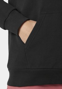 Tejido de sudadera negra con una textura suave, que presenta un bolsillo tipo canguro y puños acanalados. El dobladillo inferior es visible, contrastando con unos pantalones cortos rosas.