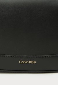 Cămașă din piele neagră cu un finisaj texturat, având inscripția aurie "Calvin Klein" și detalii subtile de cusătură. Design compact, rectangular.