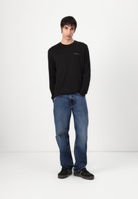 T-shirt noir à manches longues avec un petit logo sur la poitrine, associé à un jean bleu déchiré et des baskets noires. Le modèle se tient les mains dans les poches.
