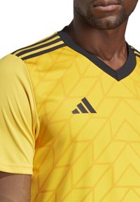 Maglia atletica gialla con motivo geometrico e texture, caratterizzata da un colletto a V nero e tre righe nere sulle spalle. Logo nero esposto.