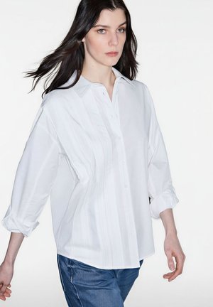 Vrouw met lang donker haar die een losvallend wit overhemd met opgerolde mouwen en een blauwe spijkerbroek draagt, tegen een effen witte achtergrond.