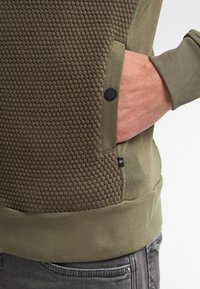 Olijfgroene sweatshirt met textuur in honingraatdesign, zijvak met knoopsluiting en ribgebreide boorden. Zachte stof met een gladde afwerking.