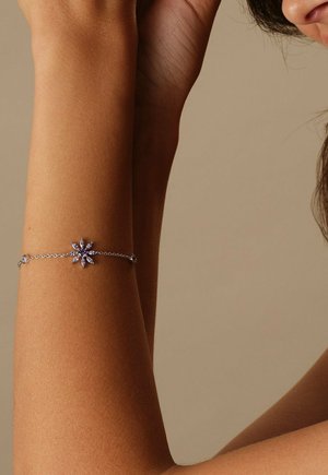 Zilveren armband met een paarse bloemvormige hanger aan een opgeheven onderarm tegen een effen beige achtergrond.