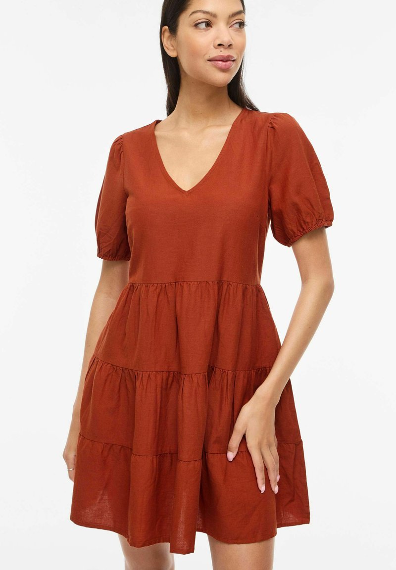 Nainen, joka pukeutunut ruosteenruskeaan kerrokselliseen mekkoon, jossa on lyhyet puffy-hihat ja V-aukko, seisoo siten, että toinen käsi koskettaa hametta.