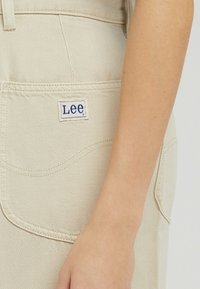 Beżowe bawełniane spodnie z tylną kieszenią, na której znajduje się niebieska metka "Lee". Materiał ma teksturowane wykończenie z widocznymi detalami szycia.